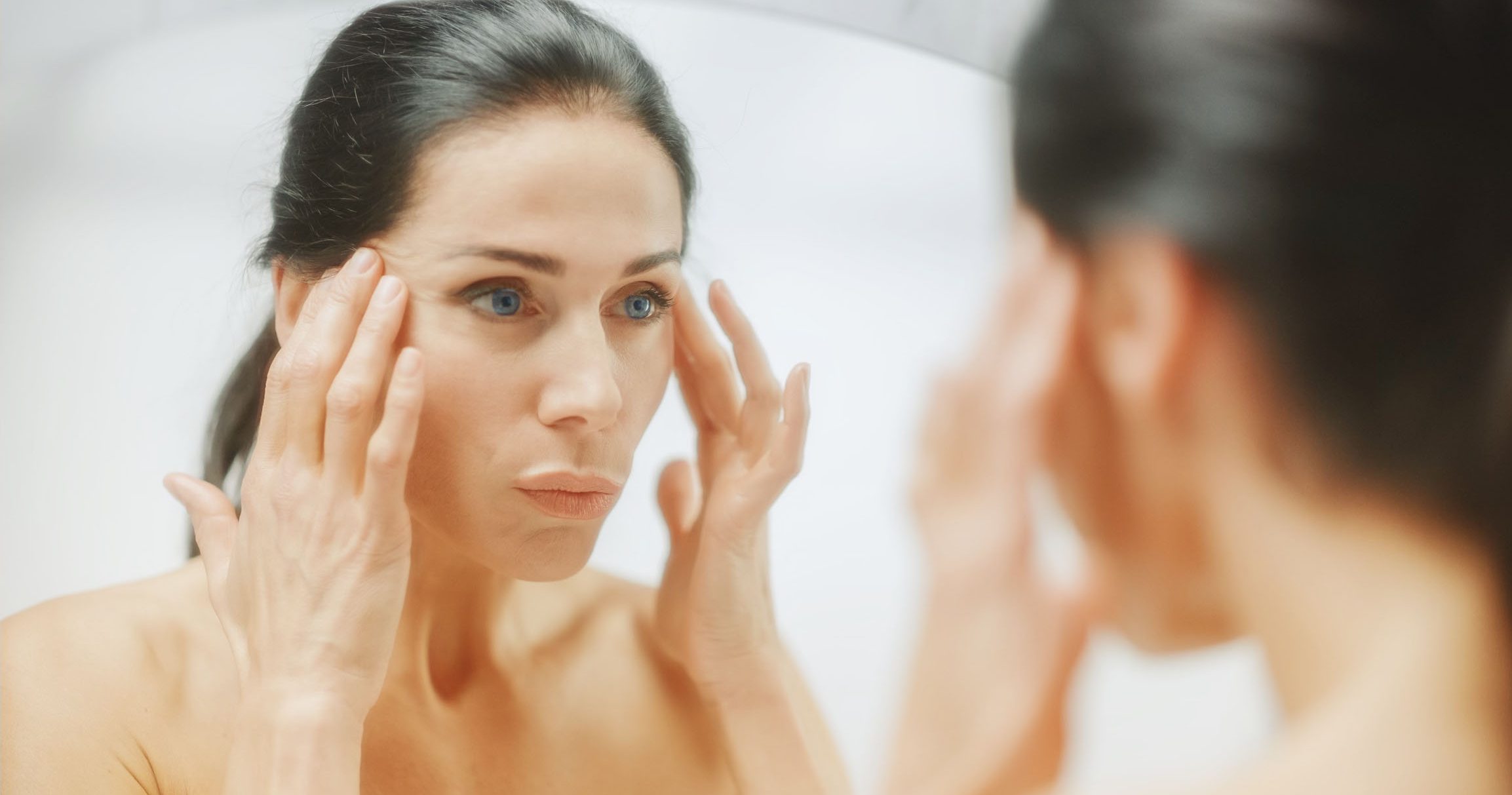 A woman in her 40s looking at herself in a mirror while considering facelift surgery.