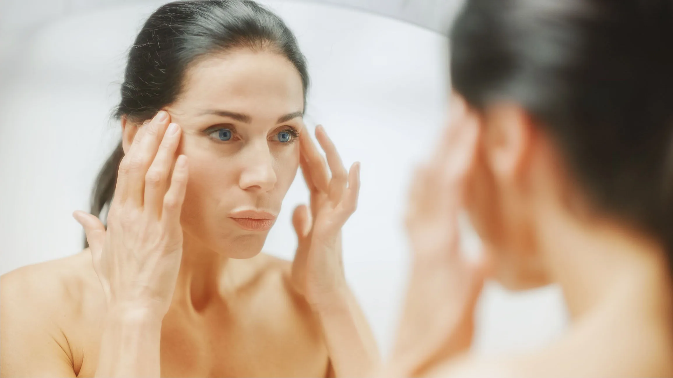 A woman in her 40s looking at herself in a mirror and thinking about facelift surgery.